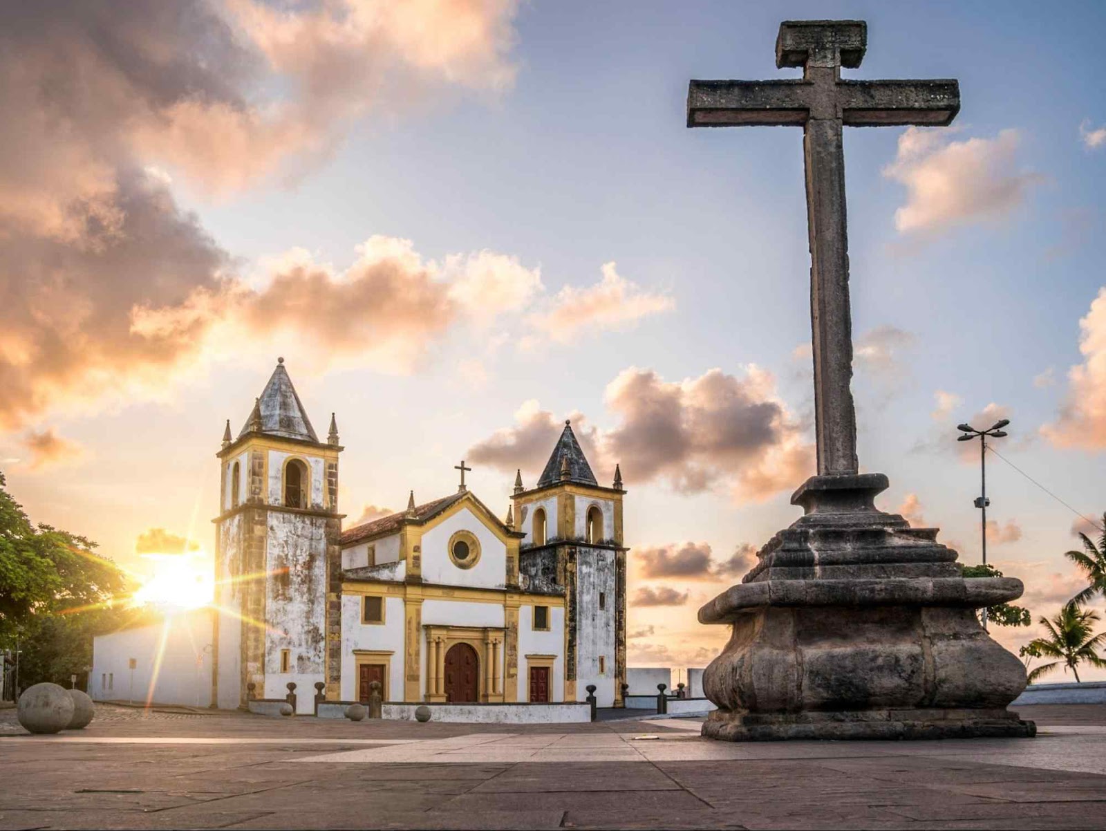 Catedral da Sé, igreja barroca de fachada branca e amarela. Ela tem duas torres com topos pontiagudos e está de frente para um grande crucifixo de pedra.