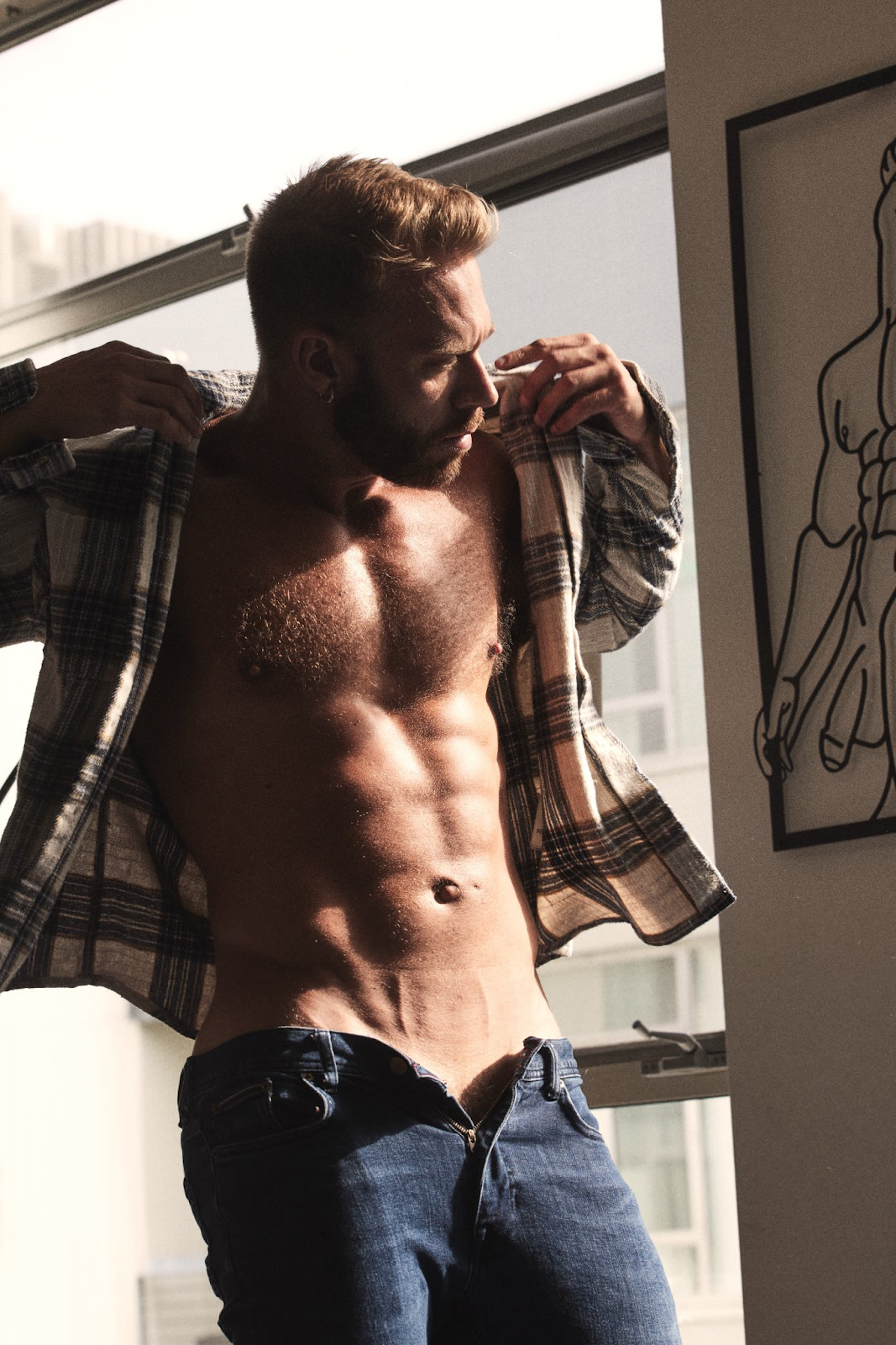 Brogan wearing an open flannel shirt staring into the distance also wearing unbuttoned blue jeans revealing the tip of his pubes