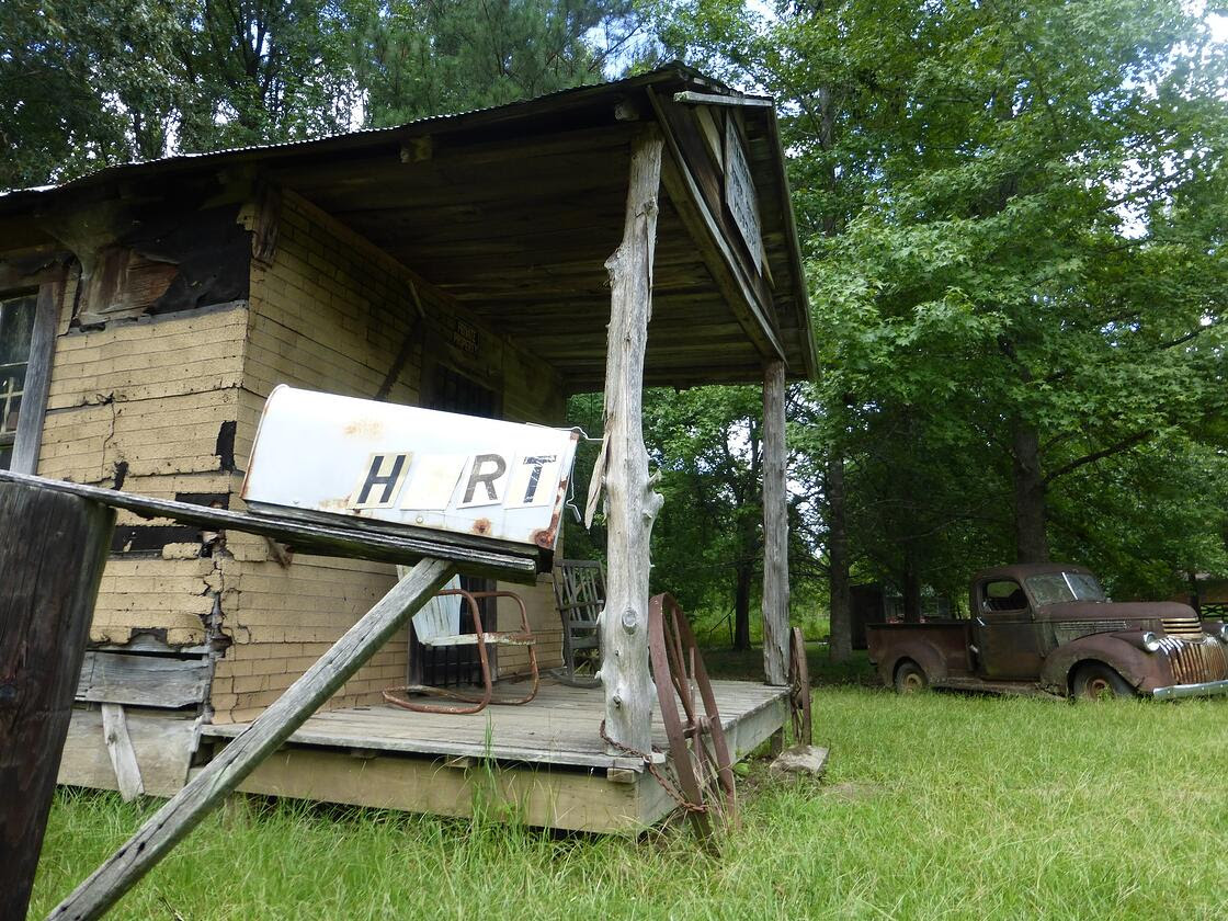 Mailbox that says “Hurt” in front of a dilapidated tan house and a rusty truck in the background. 