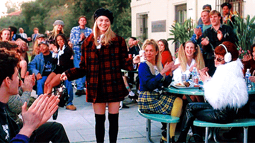 Here, the main character of 'Clueless' Cher Horowitz is seen curtsying in an outdoor school cafeteria as her fellow school peers cheer and clap at her popularity. As she curtsies she gives a smug yet cheerful look which implies "Thank you".