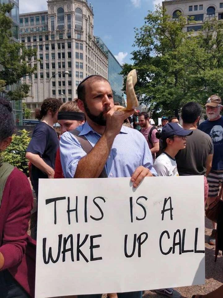 Adult person with beard and yarmulke, holding a protest sign and blowing a shofar outside in a crowd