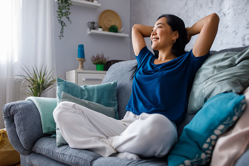 A single Asian-American woman is sitting on her couch in her living room looking at the window happily. If you are childree and childless, therapy can help you with feelings of self-confidence. Woodland Hills, CA therapy office. 91361 | 90265 | 90290 | 91367
            
