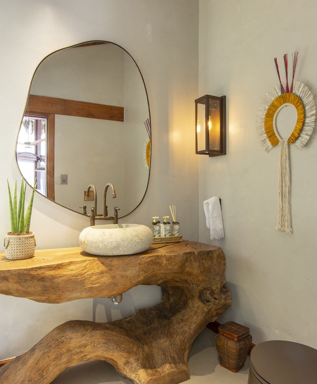 Banheiro com balcão de tronco de uma árvore, com cuba sobreposta em pedra sabão. Paredes e piso de cimento queimado com espelho orgânico, luminária na parede e enfeite indígena.
