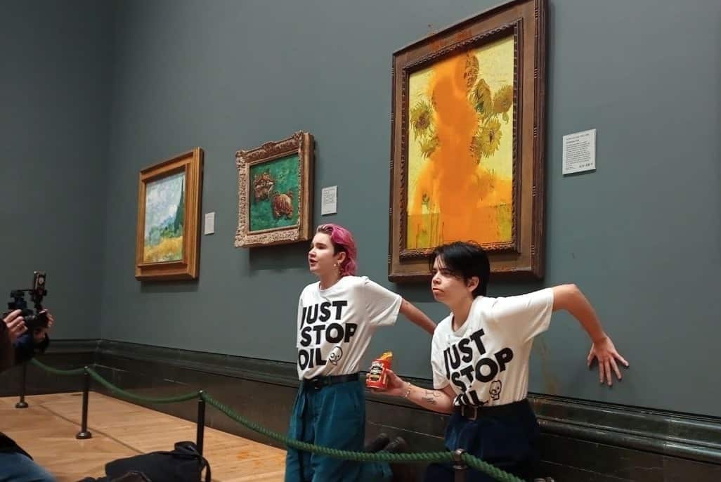 two young activist crouch by Van Gogh's Sunflowers having just thrown soup over it