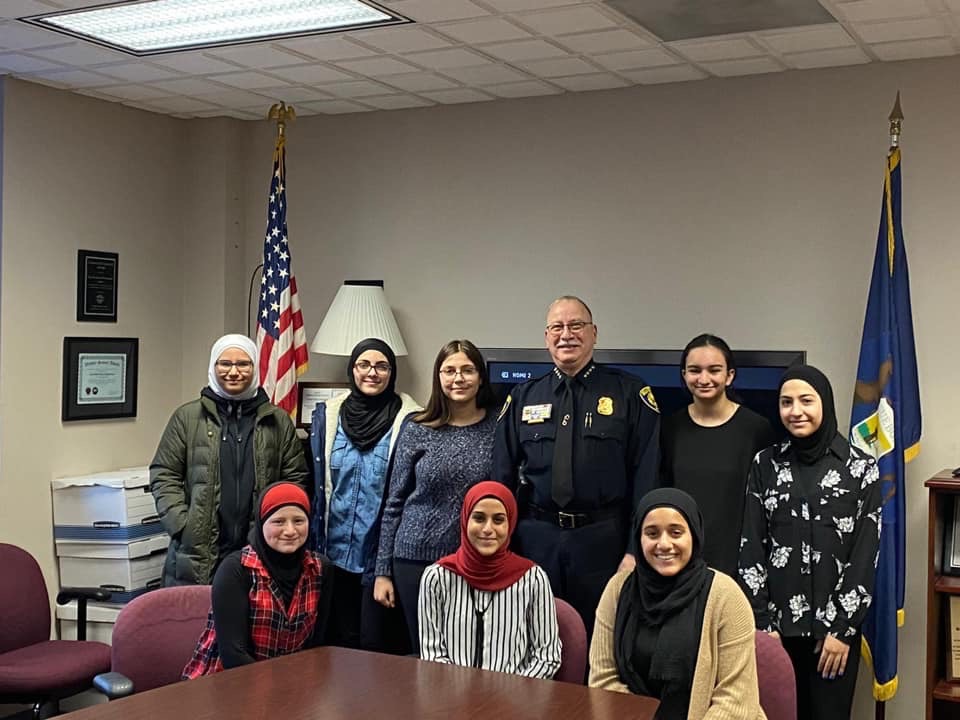 Three female students sitting at a table and five others standing with Chief Haddad in the Chief's office.