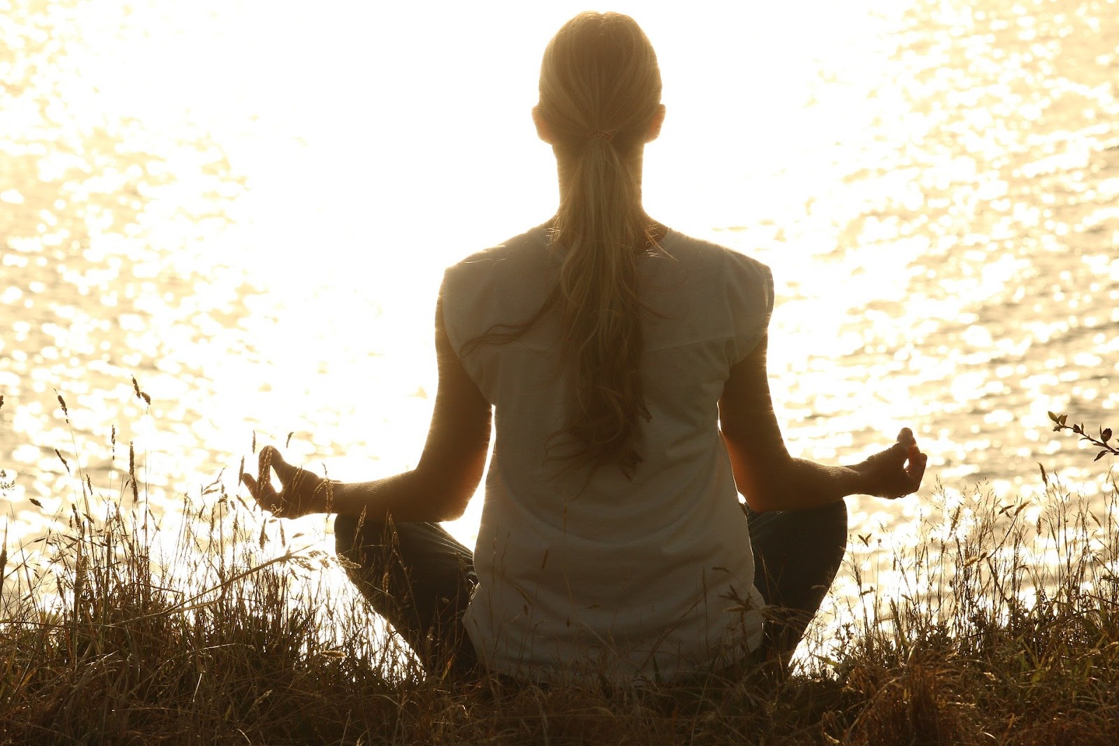 An image of a woman sitting outdoors in a meditation pose, overlooking a lack as the sun sets.