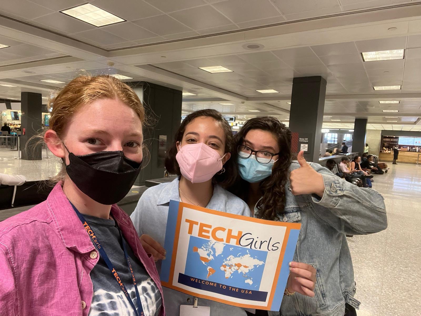 Three TechGirl Resident Assistants holding a "Welcome to the USA" sign at the airport.