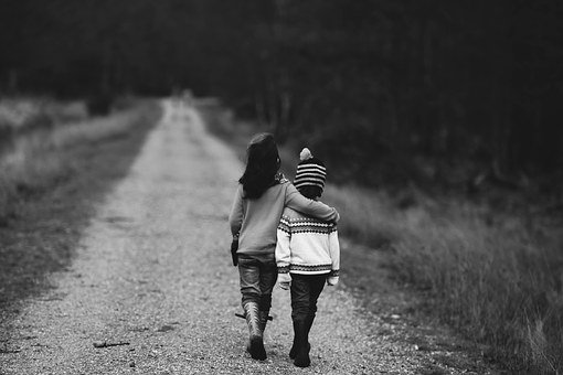 Black and white photo taken behind  two children in sweaters walking down a dirt road with their arms around each other
