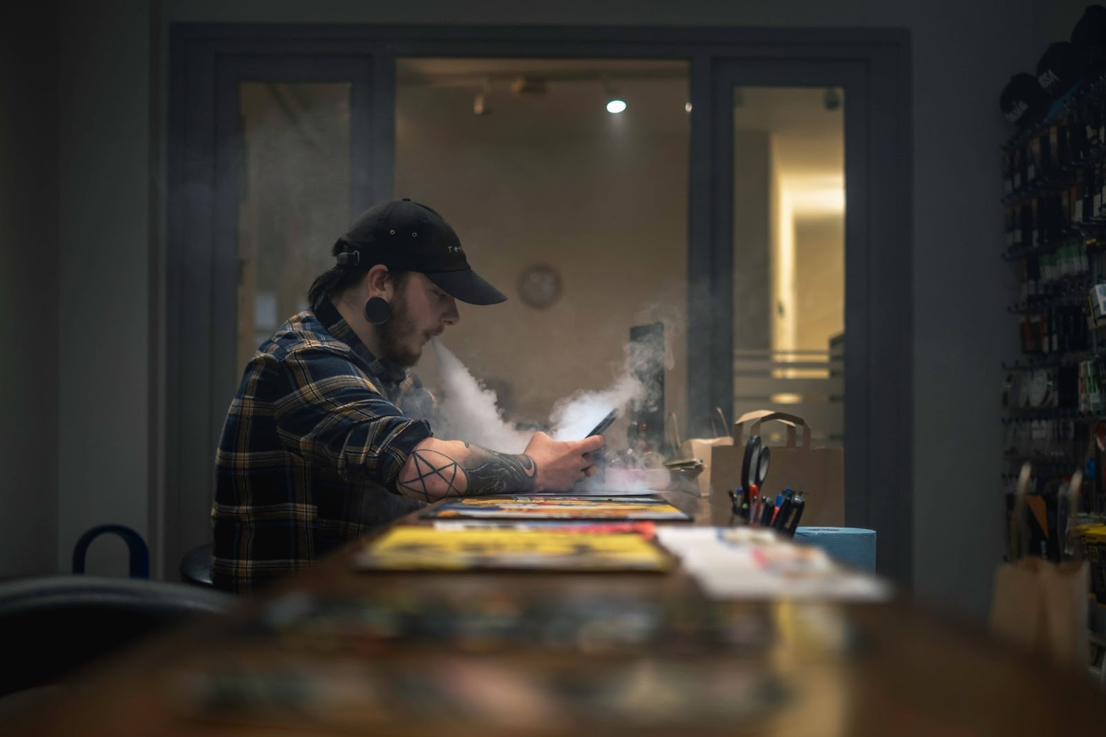 A man sat at a table on his phone and exhaling vapor from vape