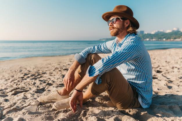 Young hipster man sitting on beach by sea on summer vacation, boho style outfit, dressed in shirt Free Photo