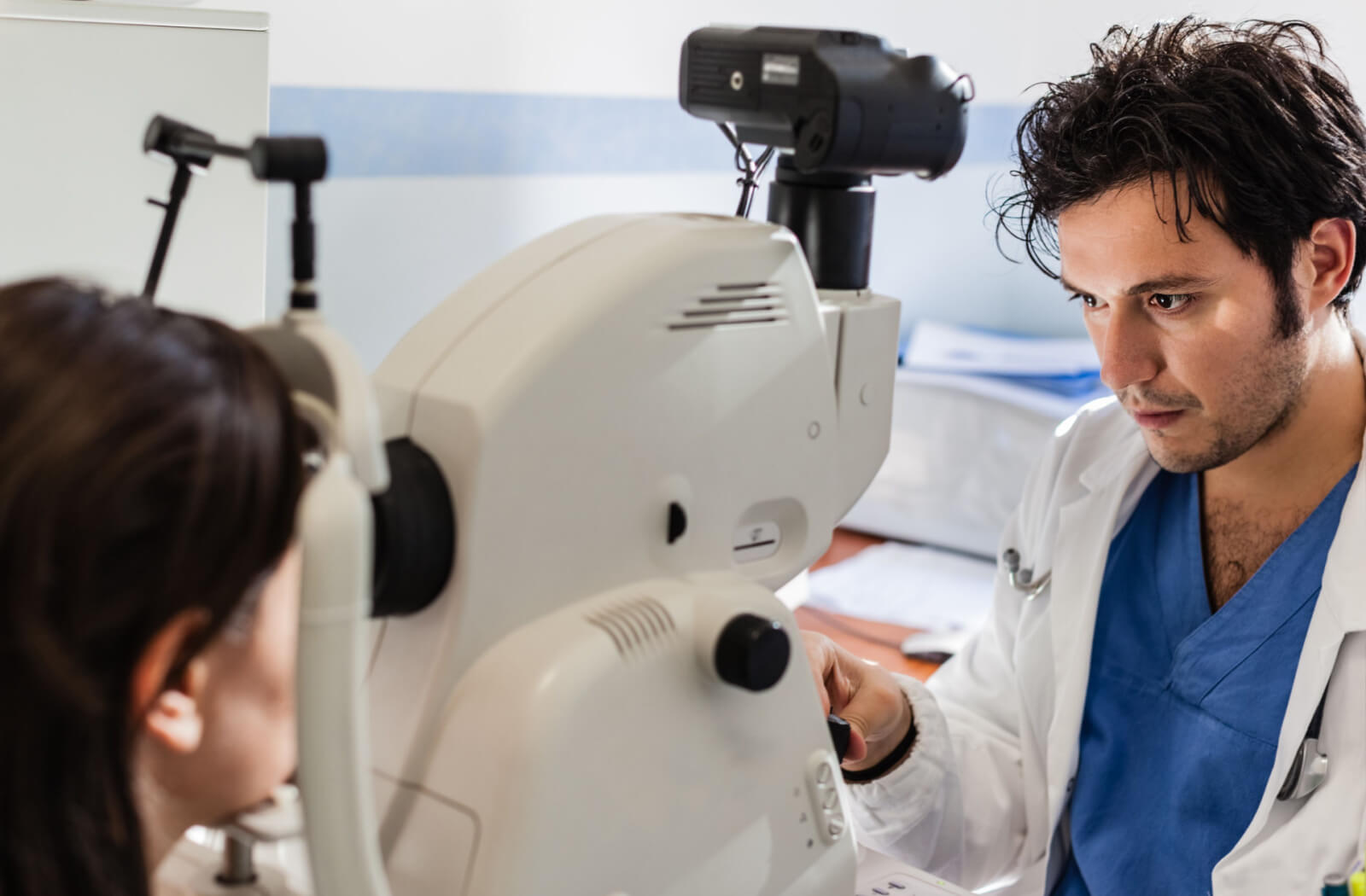 A male  optician  is operating an autorefraction machine while the patient is looking through the machine for an eye test.