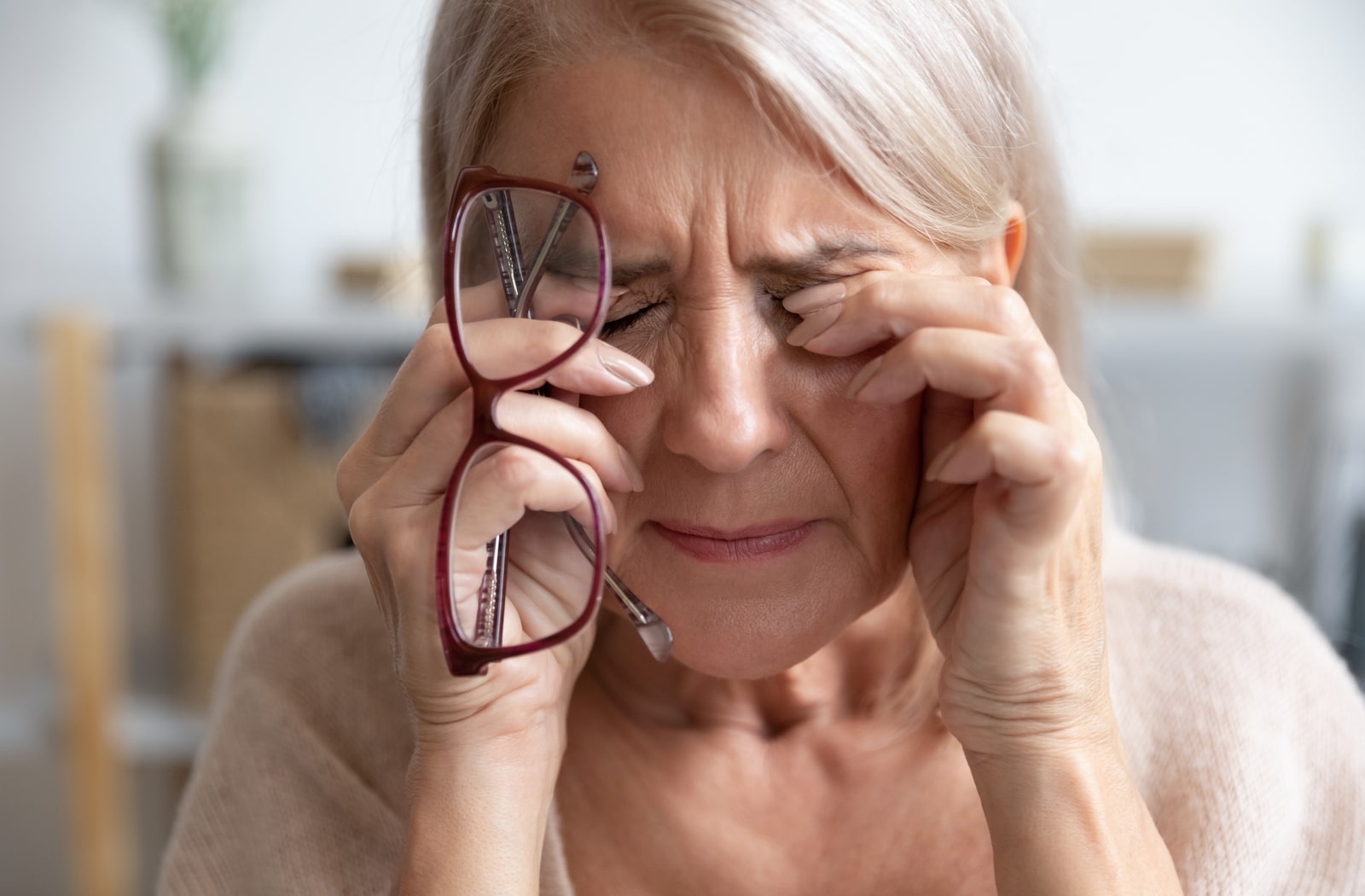  An older woman struggling with dry eye and head pain, has her hands over her eyes while close