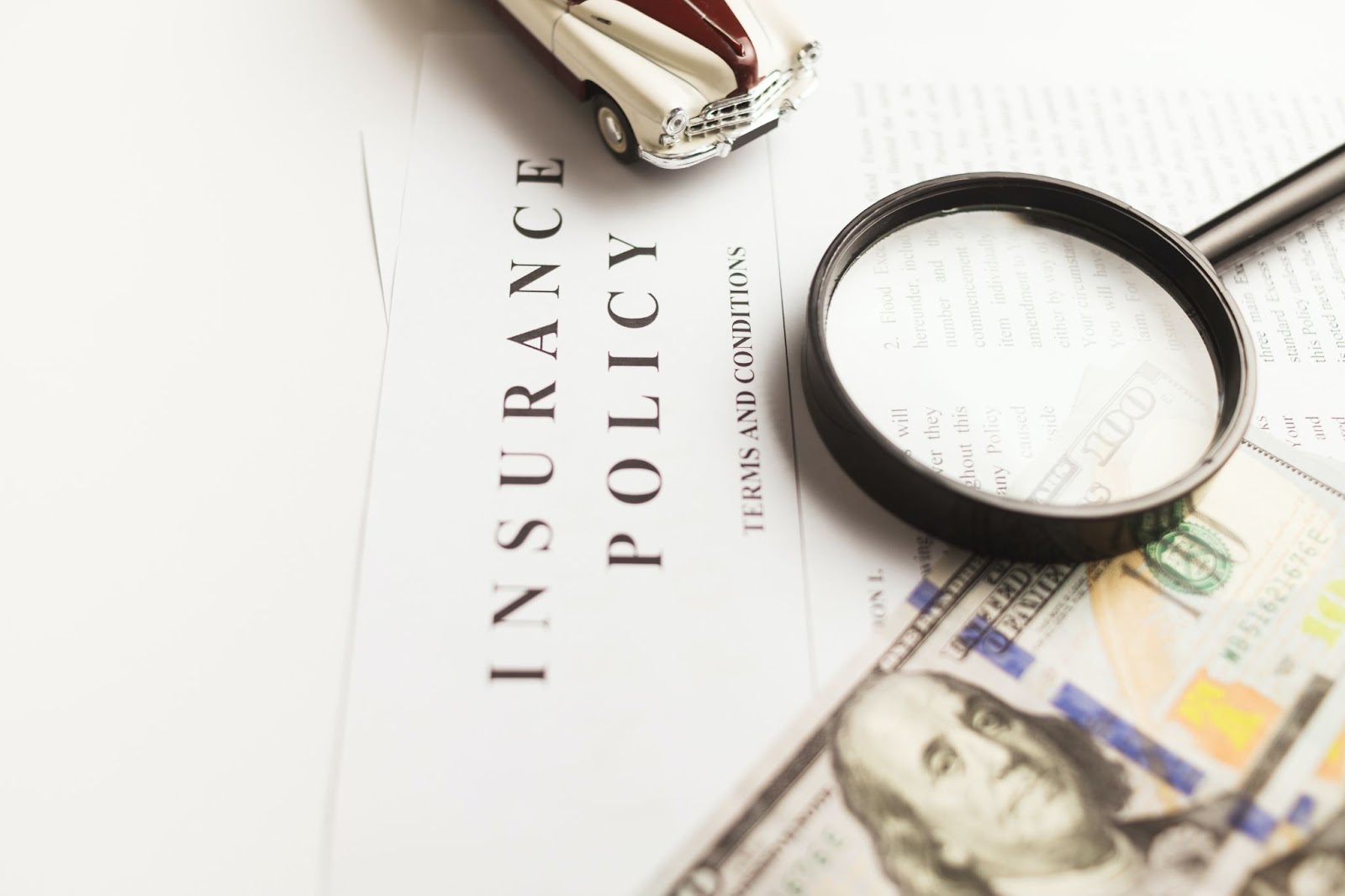 An image of an insurance policy with a magnifying glass, 100 dollar bill and a collectable toy car