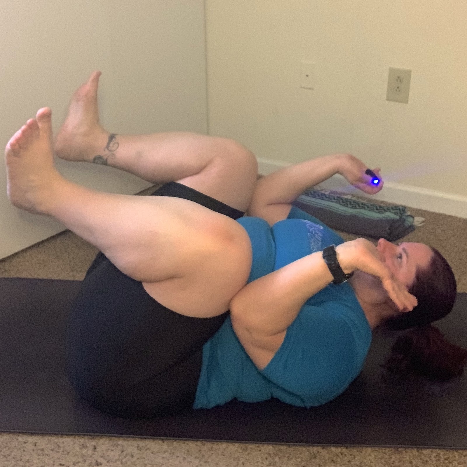 Nicole demonstrating crow pose on the back. She is laying on her back, elbows tucked on the outside of her knees, which are drawn in toward the chest. Her shoulders are slightly raised off of the purple yoga mat beneath her.