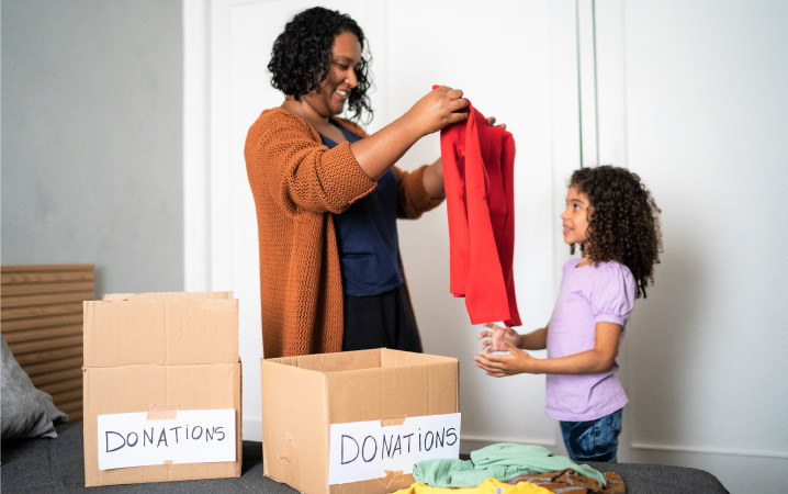 A happy mother and daughter are sorting through clothes while decluttering before their move. There are two cardboard boxes nearby labeled, “DONATIONS.”