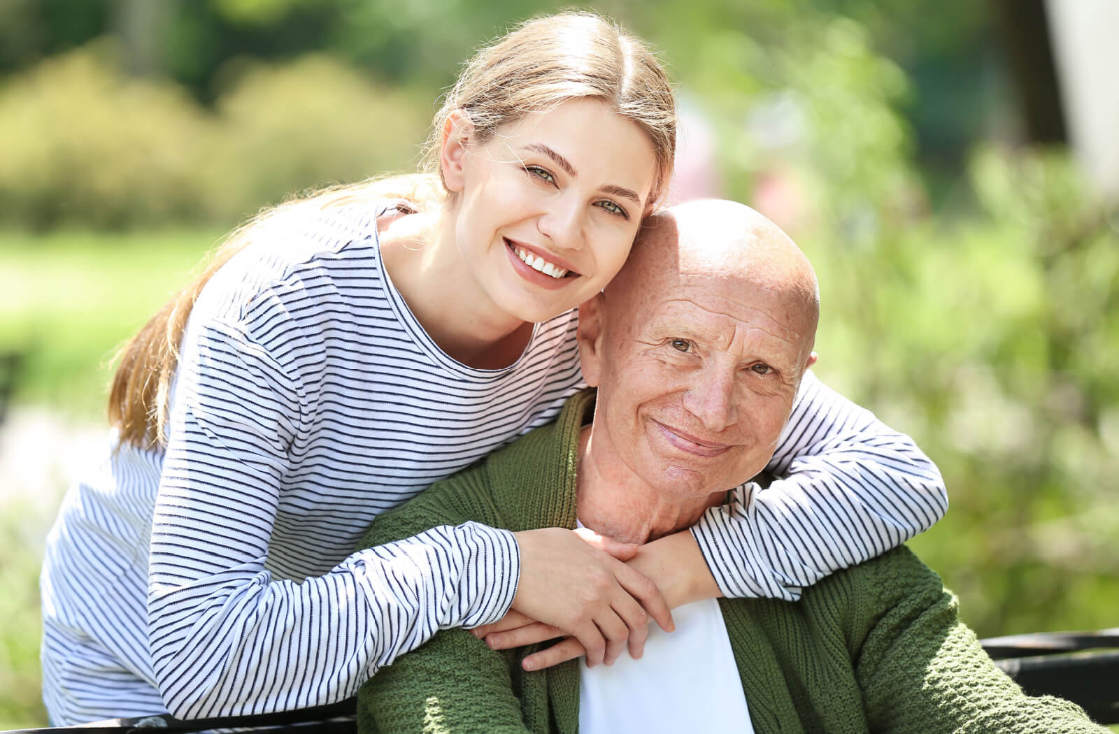 A senior man with his daughter hugging him in a park and looking directly in the camera.