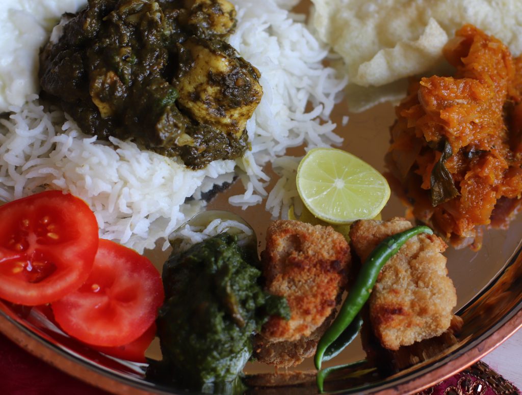 A plateful; of rice, green spinach with paneer, chicken nuggets, chutney, pumpkin, slices of tomatoes, piece of lime, crunchy papads etc , that is , rich colours and textures are seen