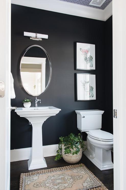 banheiro com paredes pretas, louças brancas, quadros decorativos brancos com molduras pretas, piso de madeira escura e espelho oval com moldura de inox