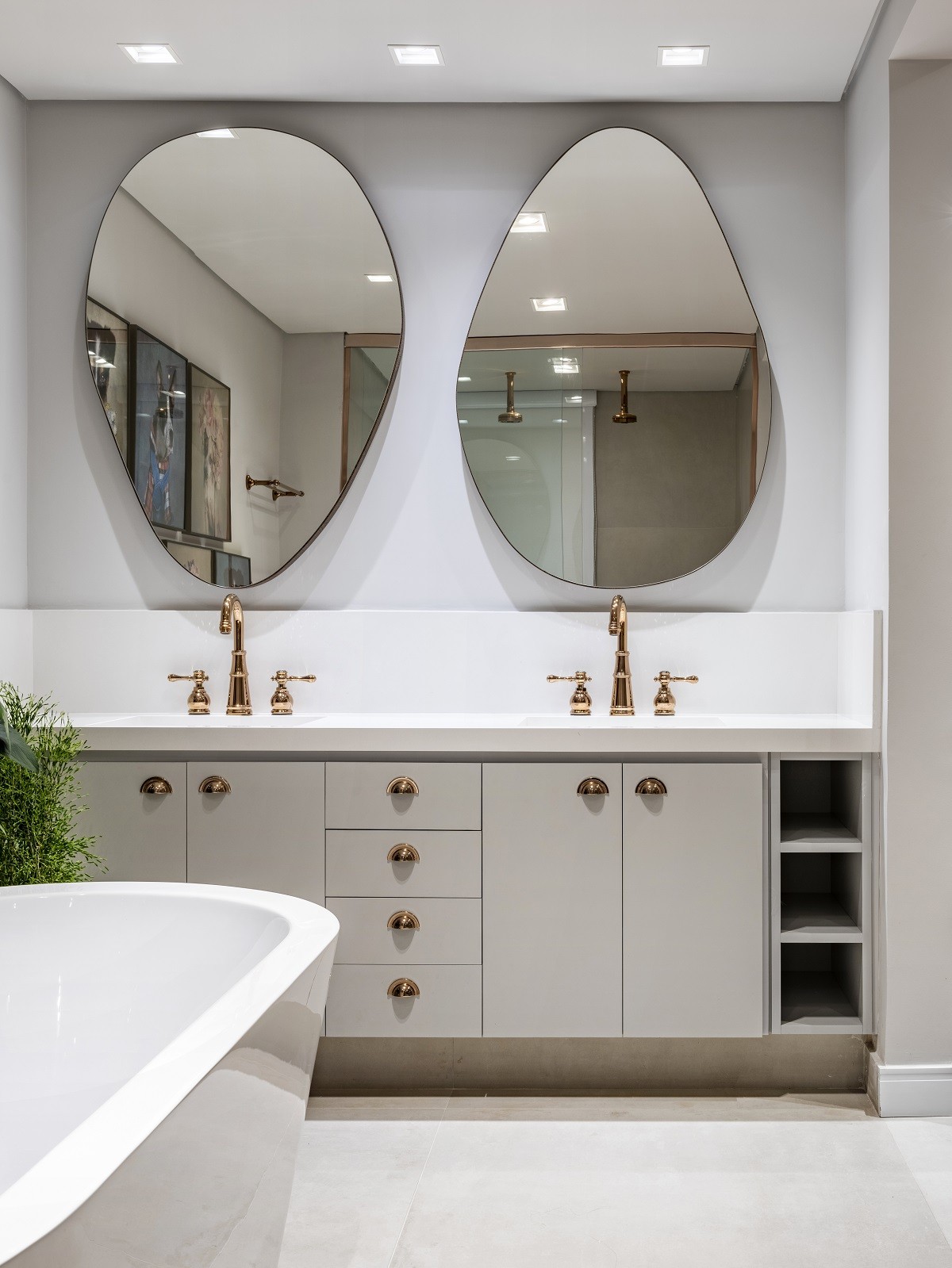 Banheiro com banheira de imersão branca, piso e paredes brancas, gabinete e bancada com duas pias com duas torneiras douradas, dois espelhos orgânicos, armário branco com puxadores dourados