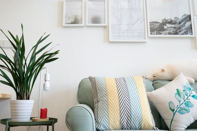 A closeup of a family's living room. There is a small green side table with a house plant and decorative lamp on it. There are pictures hung on the wall and the green couch has colorful throw pillows on it.