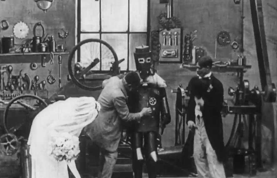 A still from The Automatic Motorist. Four people stand in a room surrounded by gears and motors, one of them is wearing something resembling a robot costume.