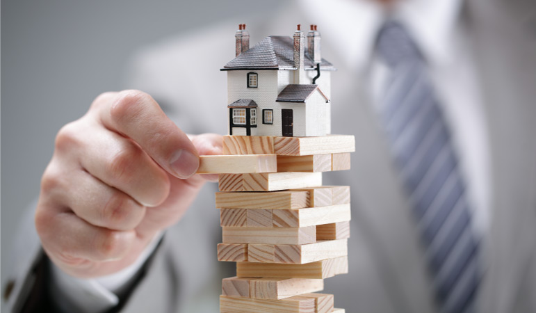 A man in business attire is pulling a wooden piece out of a Jenga tower game — except this game has a twist: A house is on top of it, symbolizing how unsteady the housing market can be.