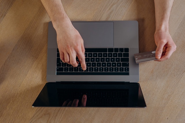 Imagem vista do alto. Mão digitando em notebook enquanto a outra segura um cartão de crédito.