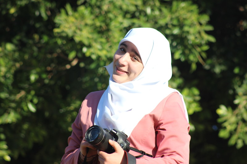 Smiling young woman holds camera while standing in front of trees