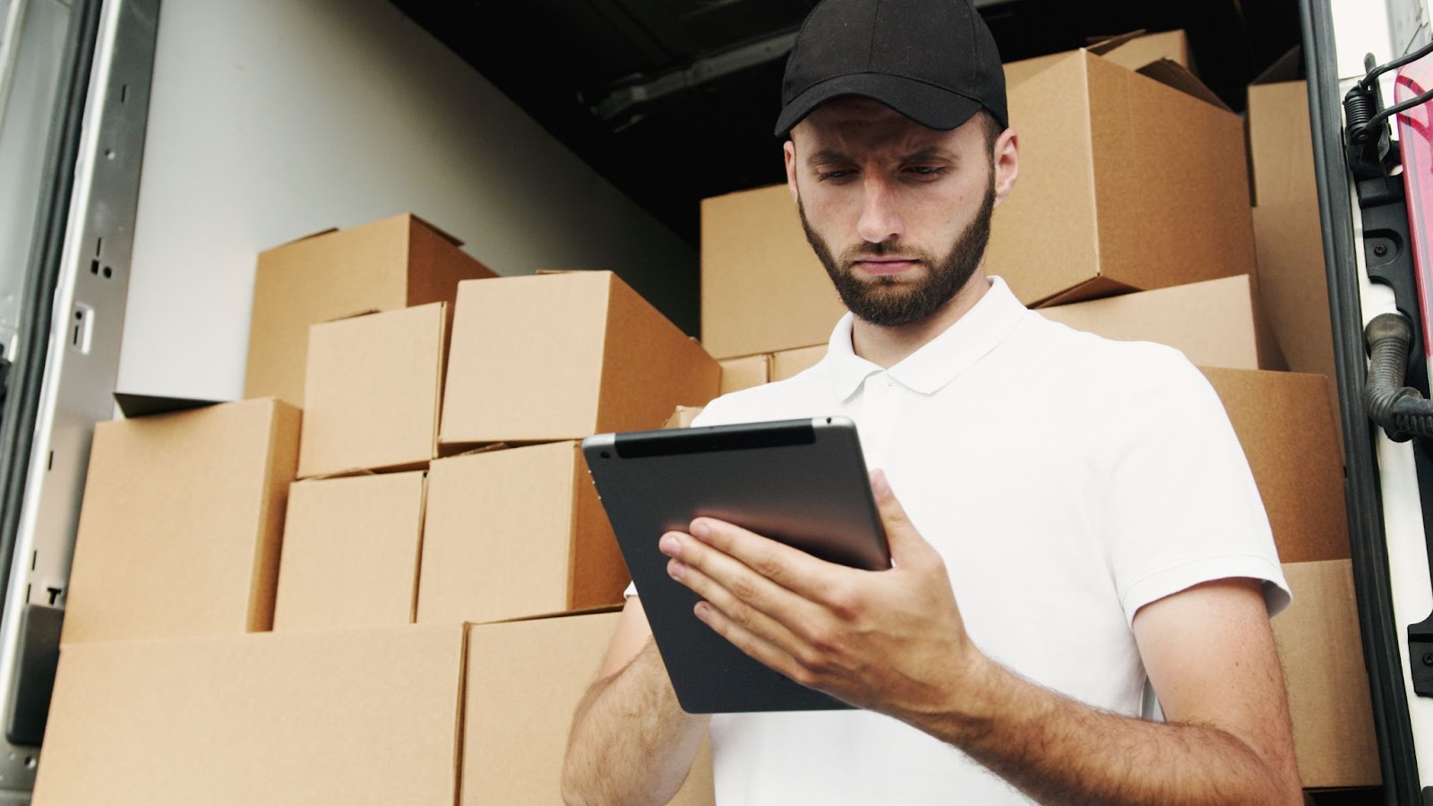 A worker looking at a tablet with brown boxes stacked in the back of his open semi truck.