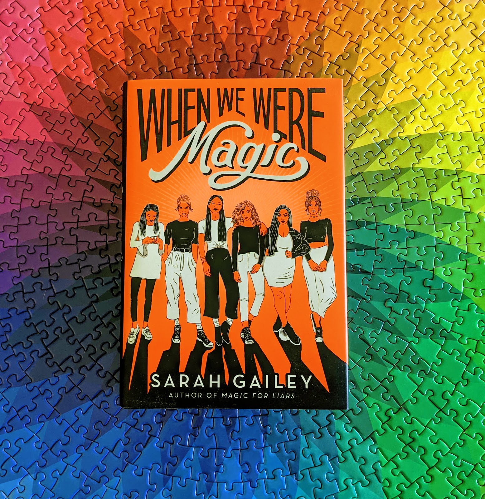 When We Were Magic, a neon orange book featuring six teen femmes in black and white clothing, resting on top of a puzzle that is a rainbow spirograph.