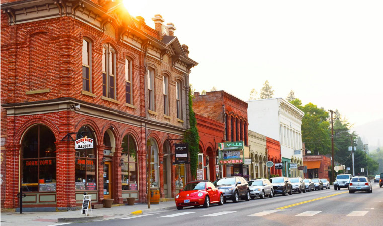 Downtown Jacksonville, Oregon, just before sunset. Cars are parked along a row of commercial shops and restaurants. Most of the buildings are made of brick and are either painted white or left a natural red color.