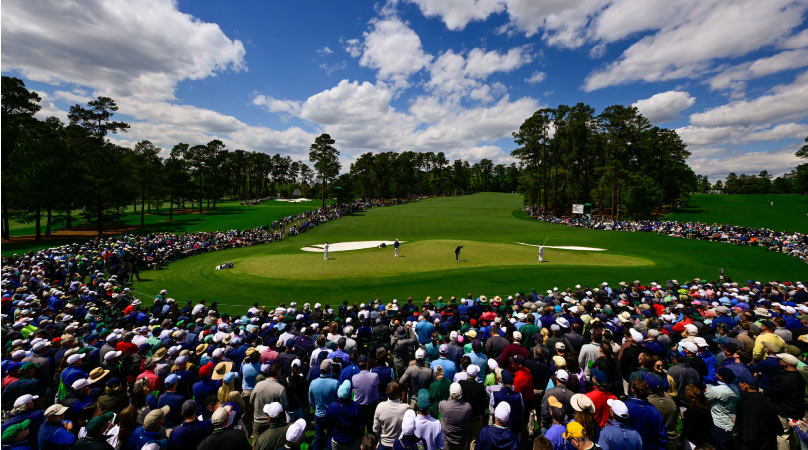 Under a bright blue sky and surrounded by crowds of spectators, professional golfers compete at the Masters Tournament in Augusta, Georgia. 