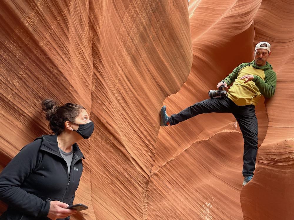 Behind the scenes portrait of photographer holding camera while straddling narrow red rock canyon and gesturing to masked second figure beneath with cell phone.
