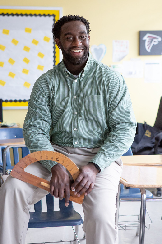 A male teacher smiles as he sits on the edge of a student desk holding a protractor in his hands.