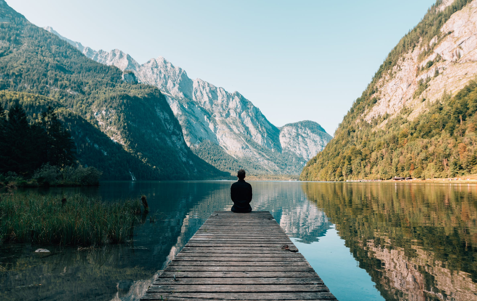 muž sediaci na móle sleduje pokojnú hladinu jazera s horami v pozadí