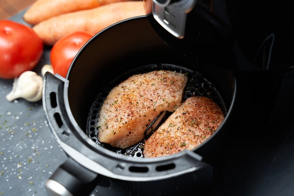 An air fryer with chicken tenderloins inside and a raw garlic, tomatoes, and carrots on the side