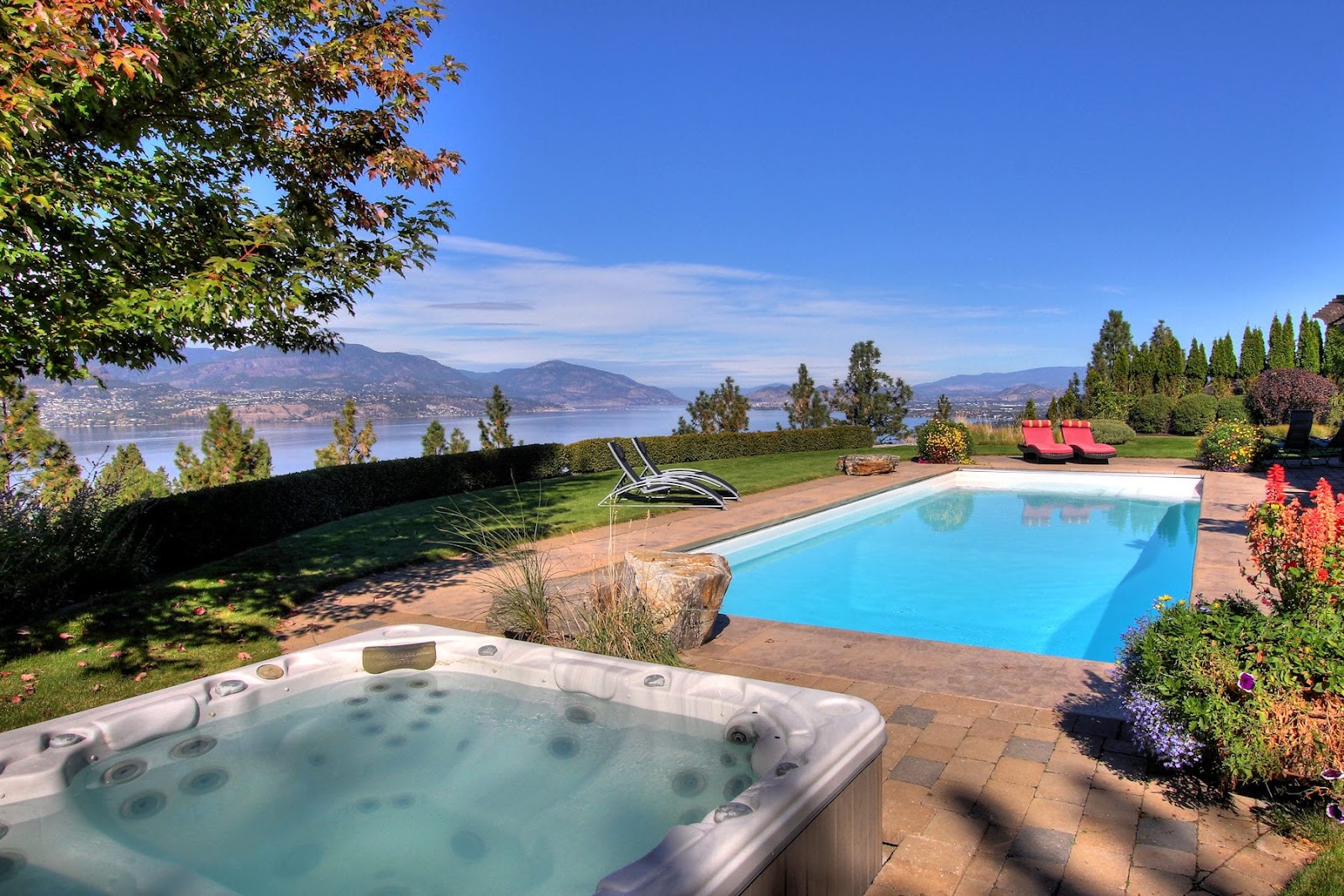 A hot tub and pool side by side on a property set on the hillside with a view of Okanagan Lake in the background.