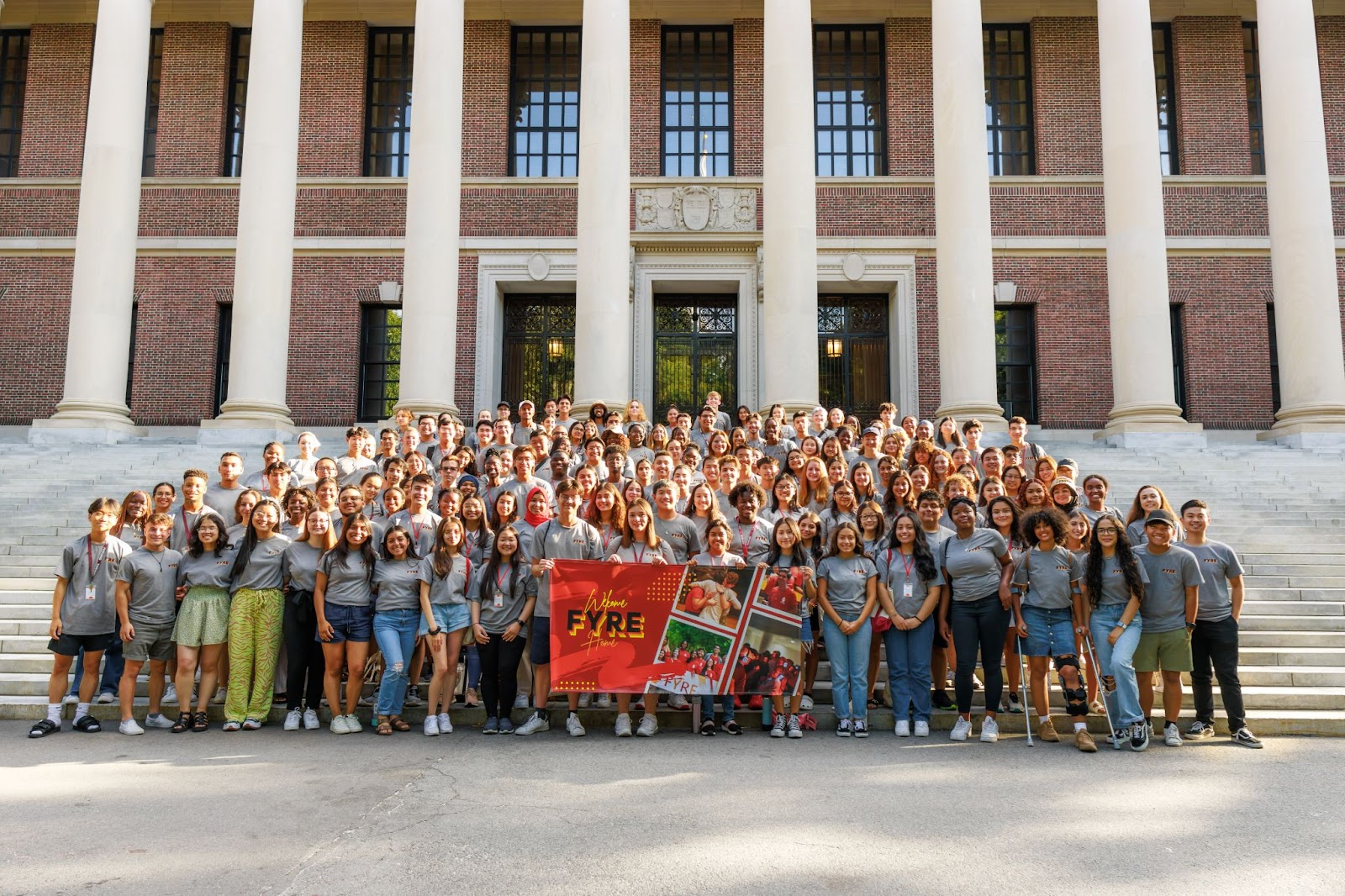 A group of 120 first years stand in front of the steps of Widener Library for a photo of their First Year Retreat Experience cohort. They hold us a FYRE banner and wear their FYRE shirts. 
