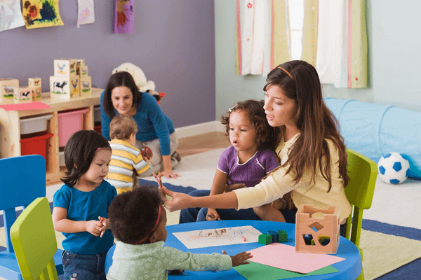 A teacher sits at a table with two toddlers sitting across from her and one on her lap. She holds out two orange crayons to one of the children. In the background of the photo, another adult is playing with a toddler as they hold a toy. 
