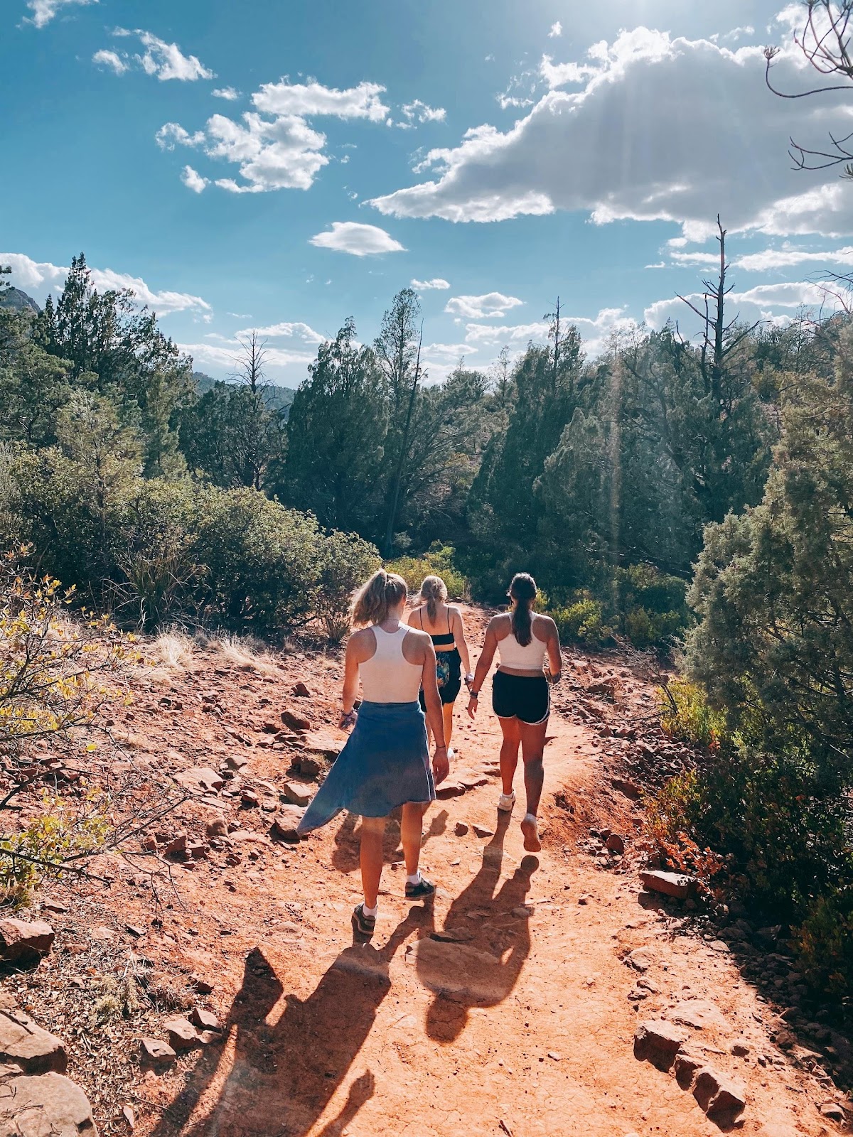 three young women hiking the Grand Canyon in shorts, tank tops, and hats. The sky is blue with a few white clouds.