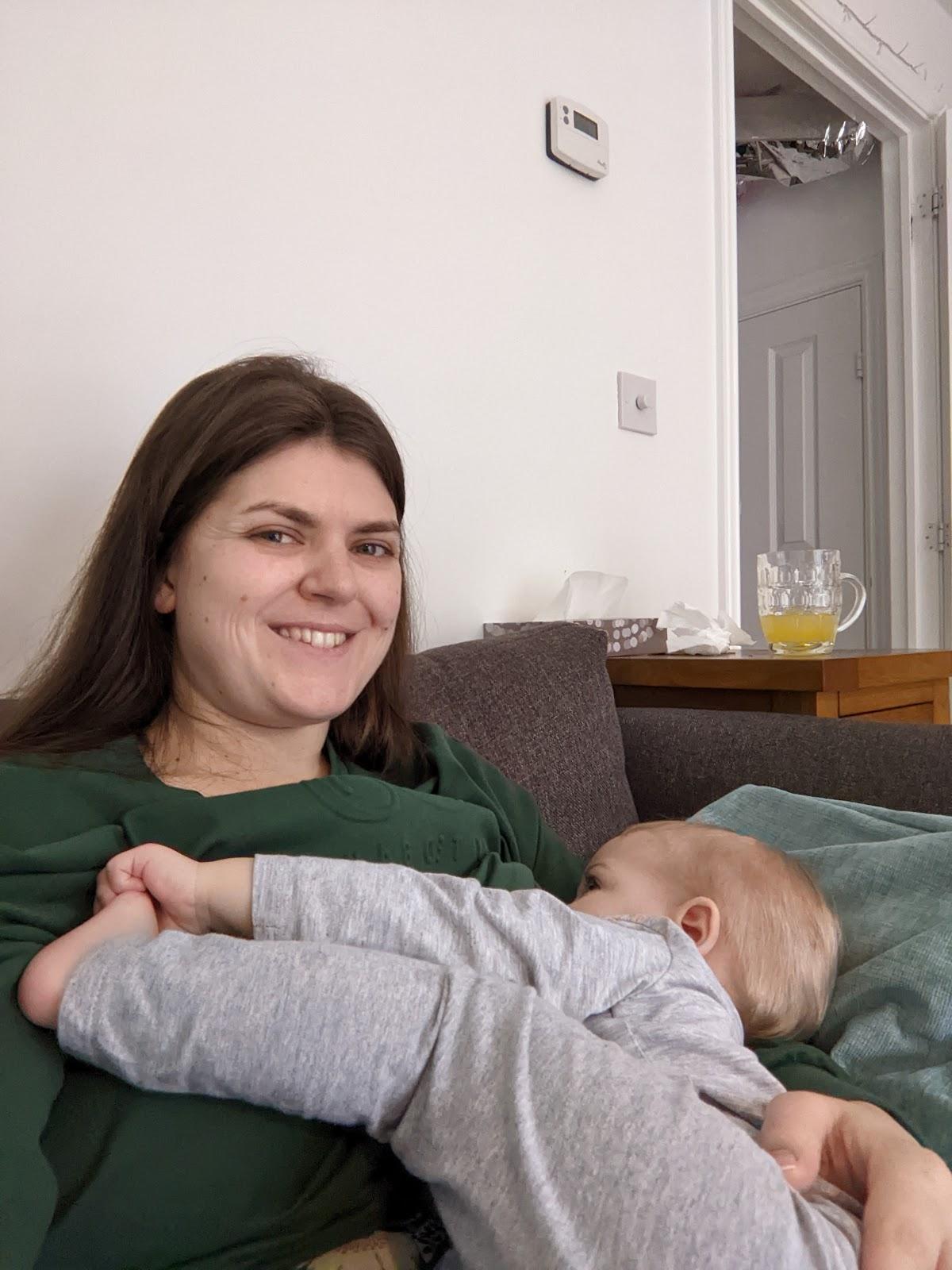 Fiona is sitting on a grey sofa, wearing a dark green top and feeding her daughter who is holding her left leg up with her left hand while feeding. Her daughter is dressed in grey.