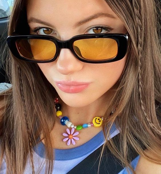#DescriçãoDaImagem: foco da imagem no rosto de mulher de cabelos castanhos que usa óculos preto com lentes laranjas, blusa azul e colar colorido. Foto: Pinterest.