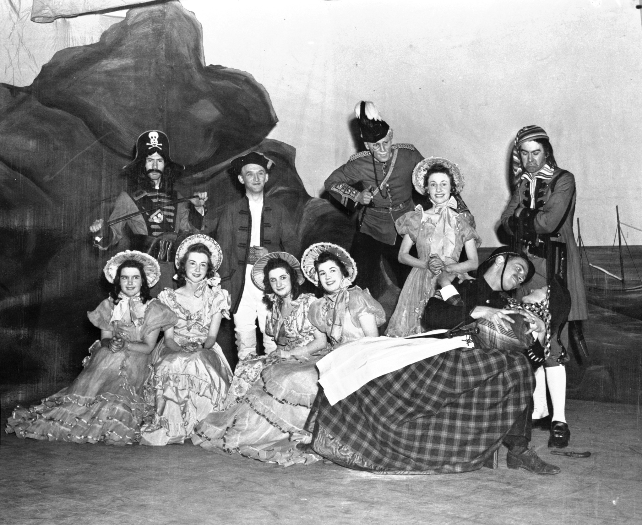 Black and white photo. Two rows of performers pose for the camera on stage. A rock and ship's masts are painted behind them. The men wear military uniforms and the women wear long frilly dresses with bonnets.