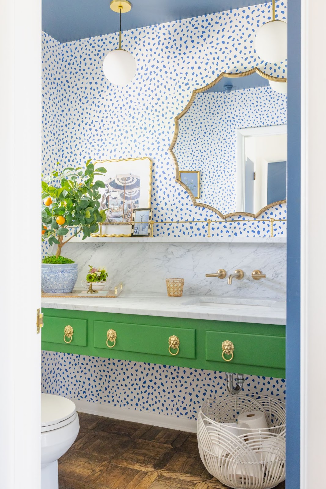 A colorful bathroom features a boldly painted green vanity cabinet, blue and white print wallpaper with touches of gold and polished brass as accent pieces