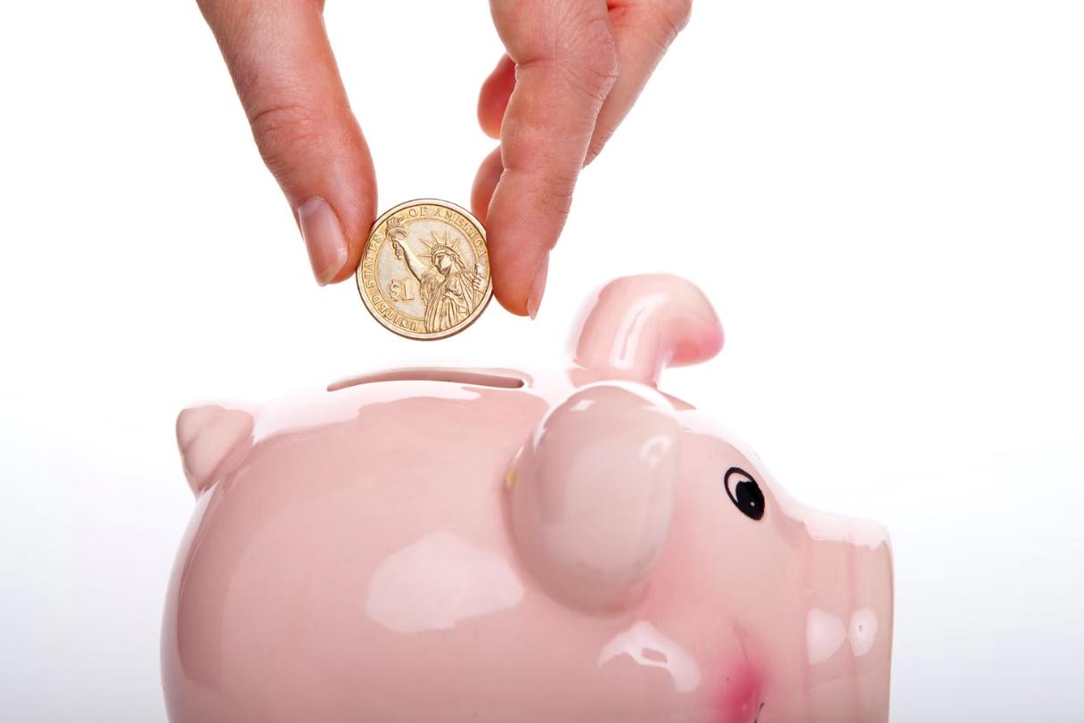 A hand holding a coin over a piggy bankDescription automatically generated with medium confidence