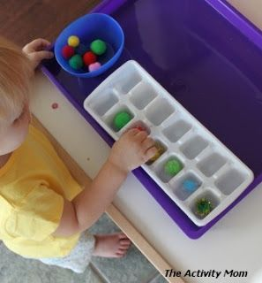 A child in a yellow shirt standing in front of white ice tray and a blue bin filled with pom poms on a purple tray. The child is placing one pom pom in each section of the ice tray.