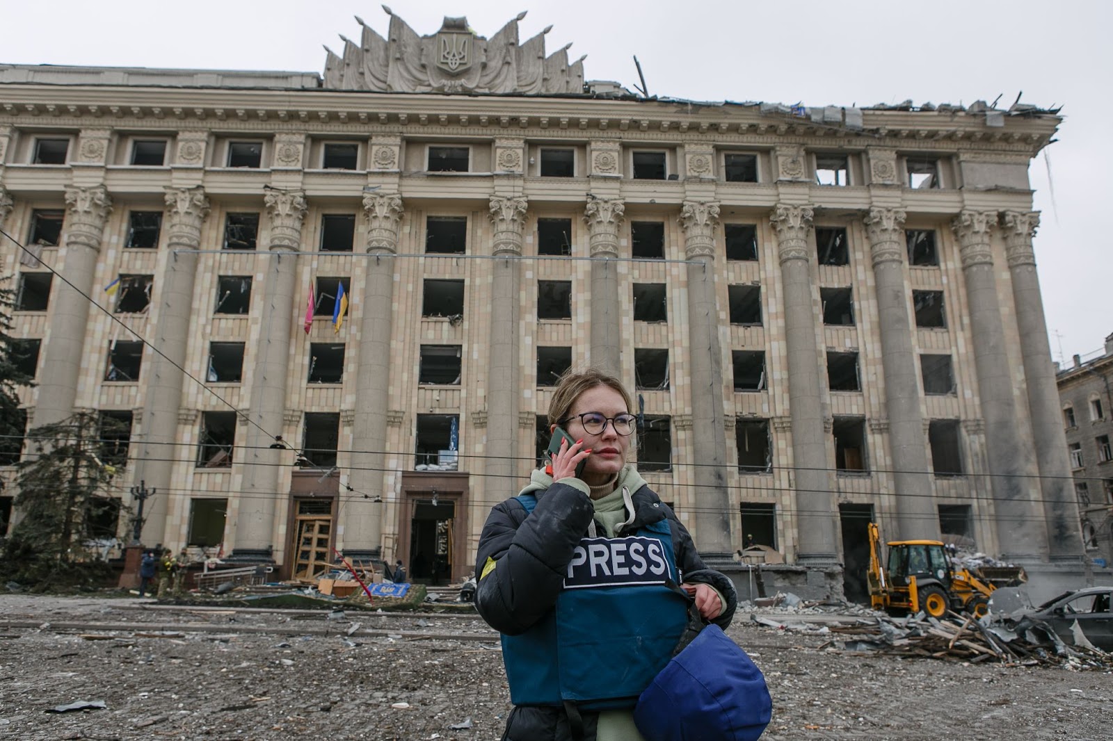 Journalist with bulletproof vest talking on the phone in front of a Ukrainian building struck by Russian attacks.