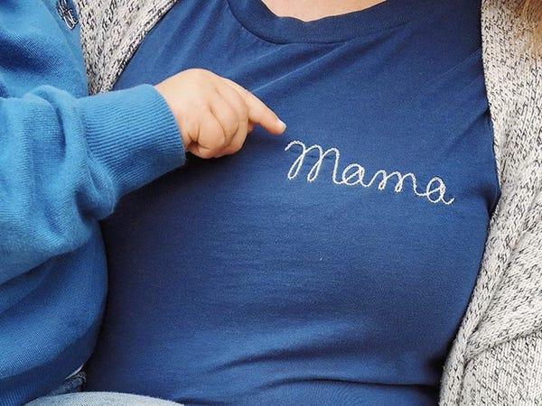 person wearing blue t-shirt with cursive embroidery that reads "mama" and baby's hand pointing at text