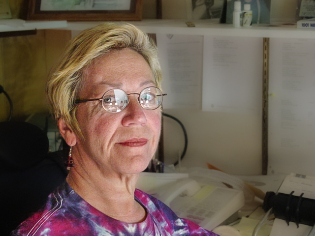 Blonde woman with round glasses and tie-dye shirt looks stoically at the camera. 