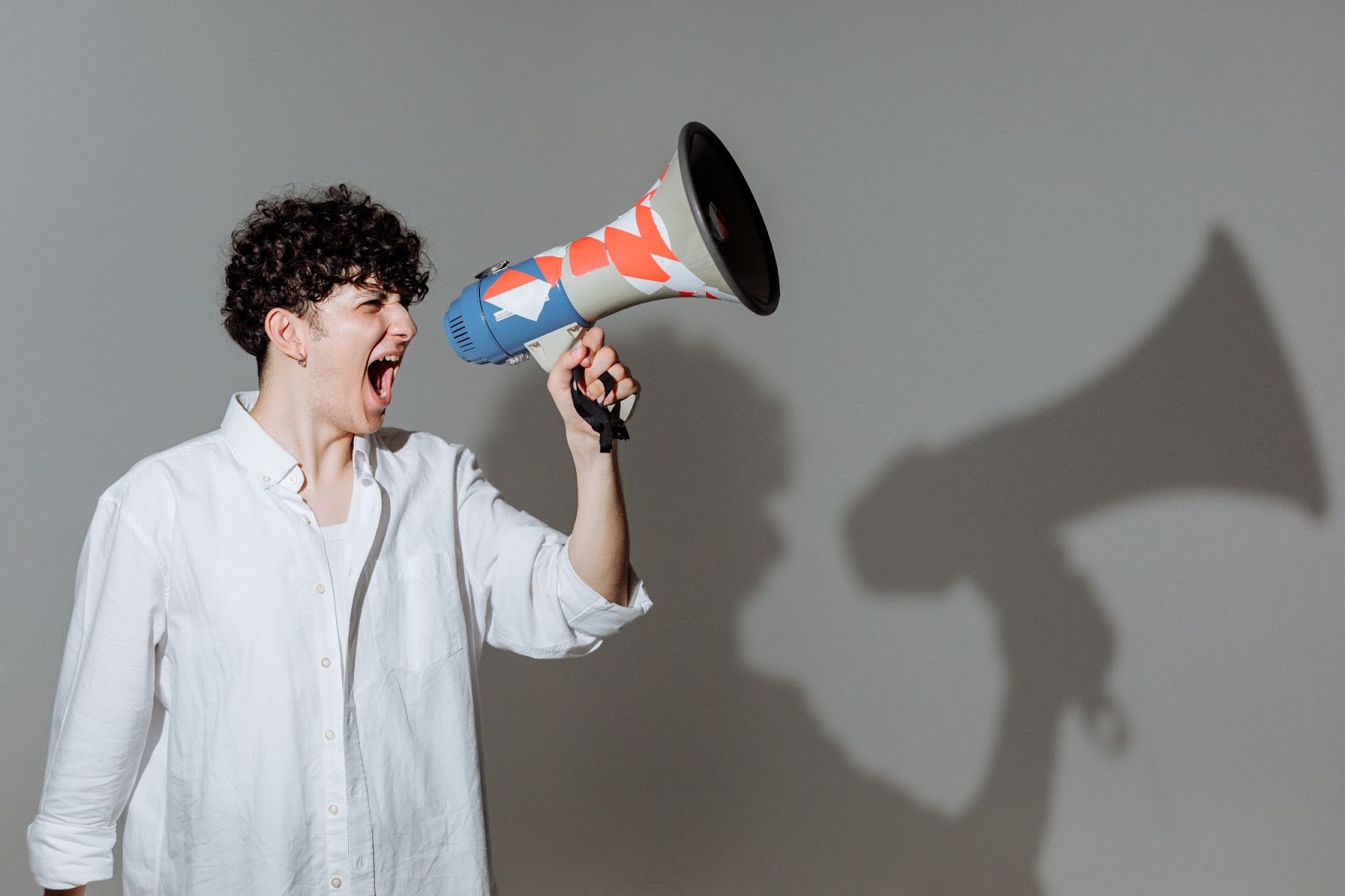 Someone stood in a white shirt, with short curly hair, shouting into a megaphone. Their shadow is prominent behind them. 